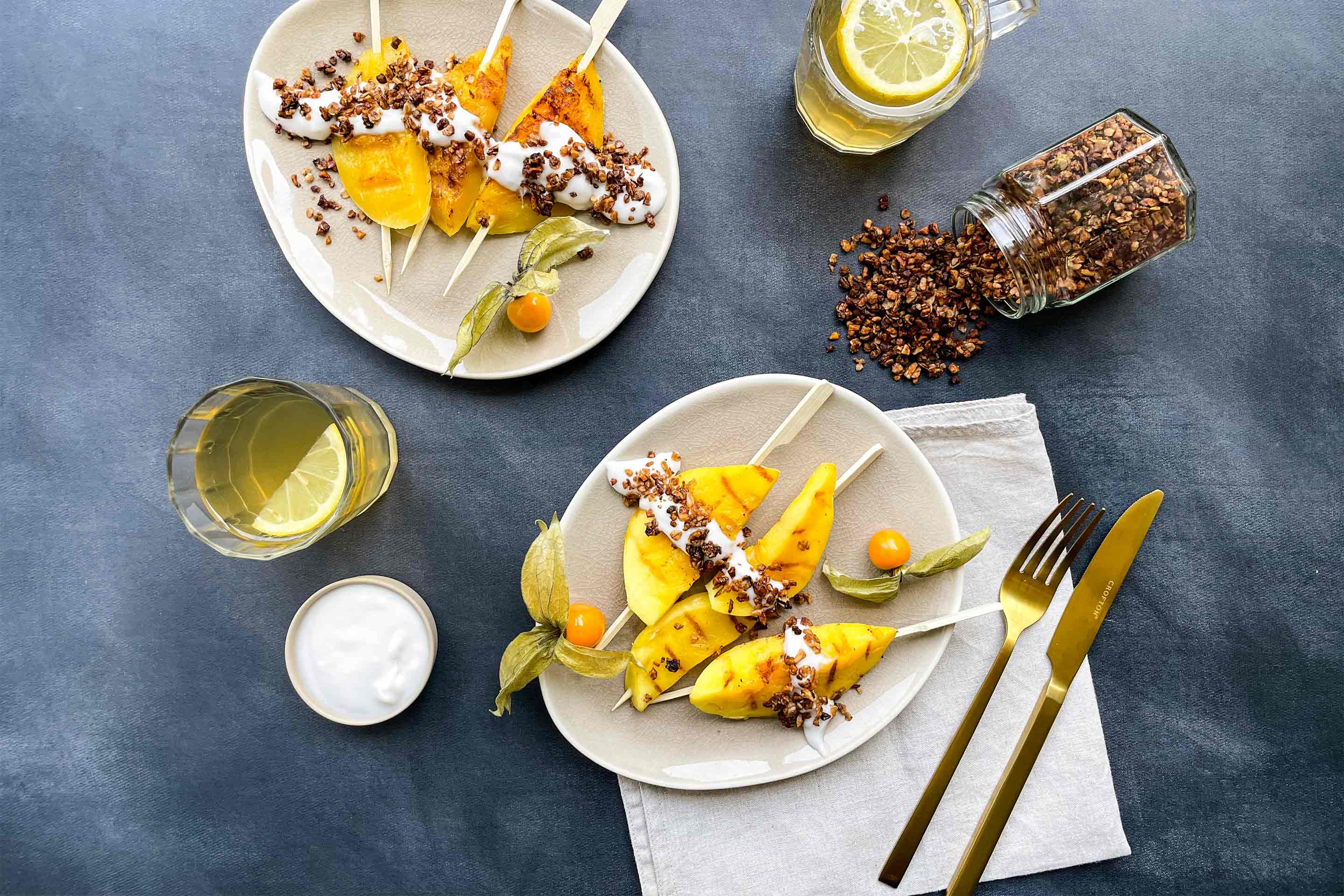 Аранжировка на плодови шишчета с манго, буркан с домашна гранола изсипан върху масата и лимонада в чаши