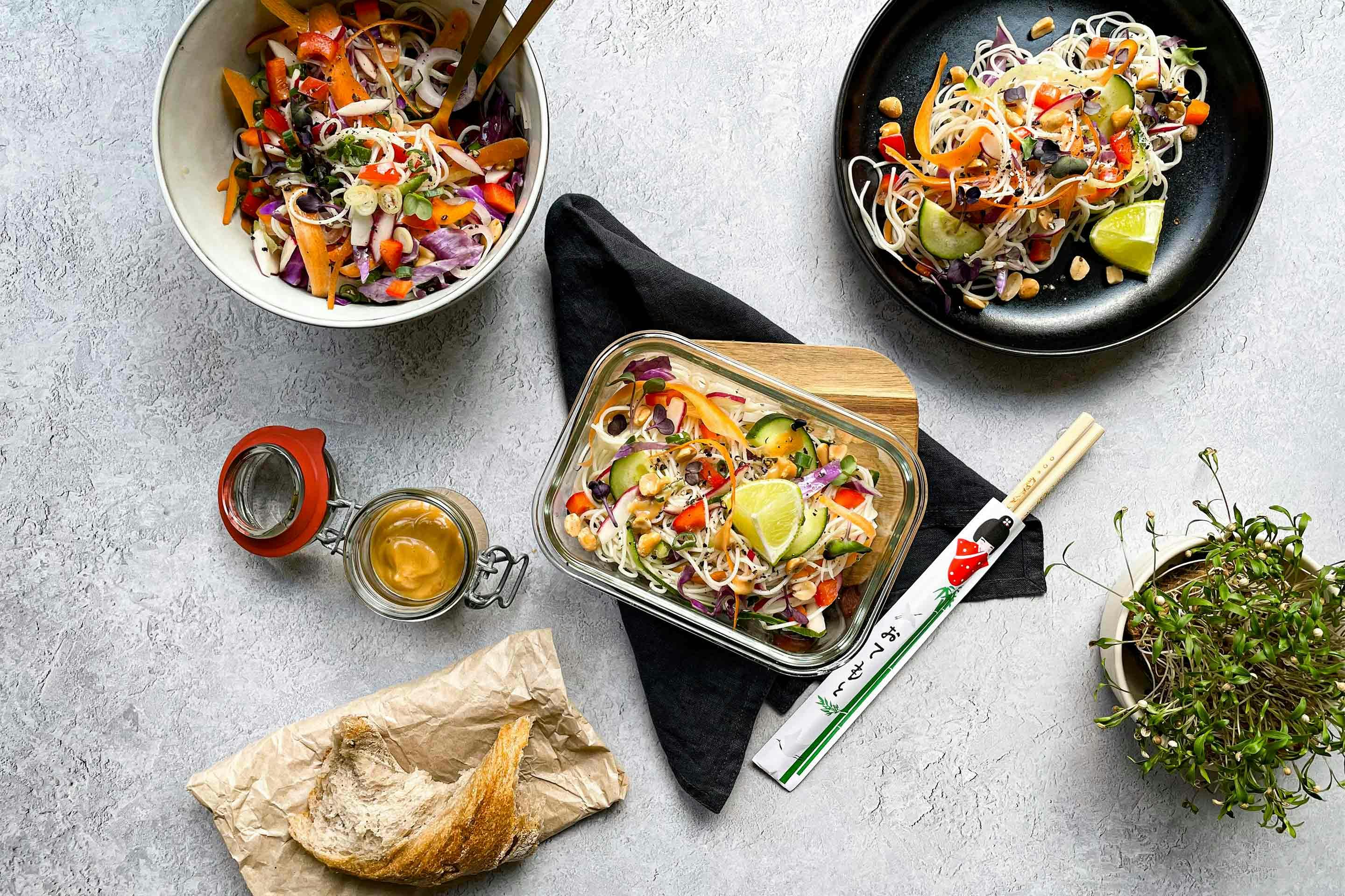 Три порции салата с нудли и зеленчуци поднесени в купичка, чиния и кутия за съхранение, късче хляб и бурканче с оранжев сос отляво, малка саксия с кълнове отдясно