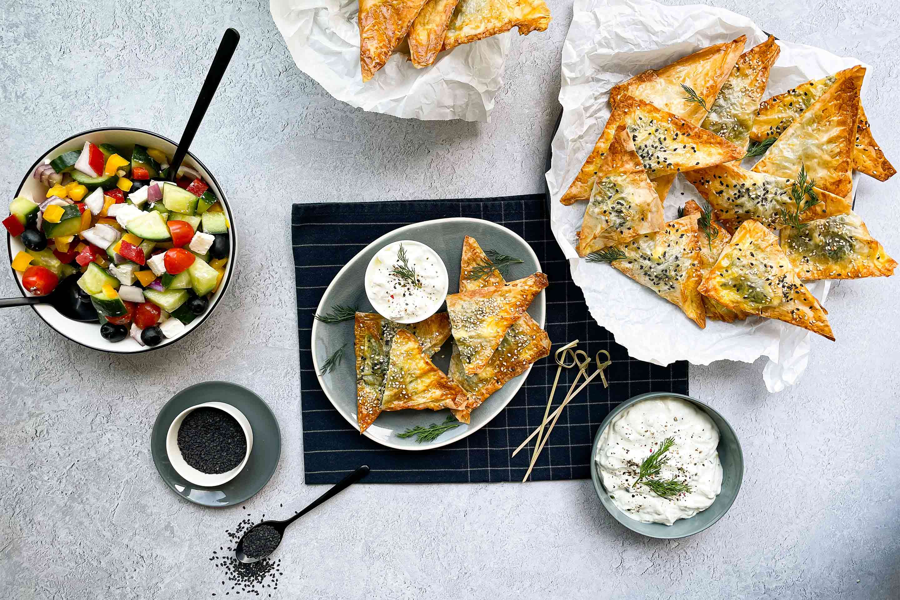 Гръцка баница със спанак на триъгълничета в чиния върху малка, черна покривка, поднесена със стръкчета копър, чеснов сос и гръцка салата.
