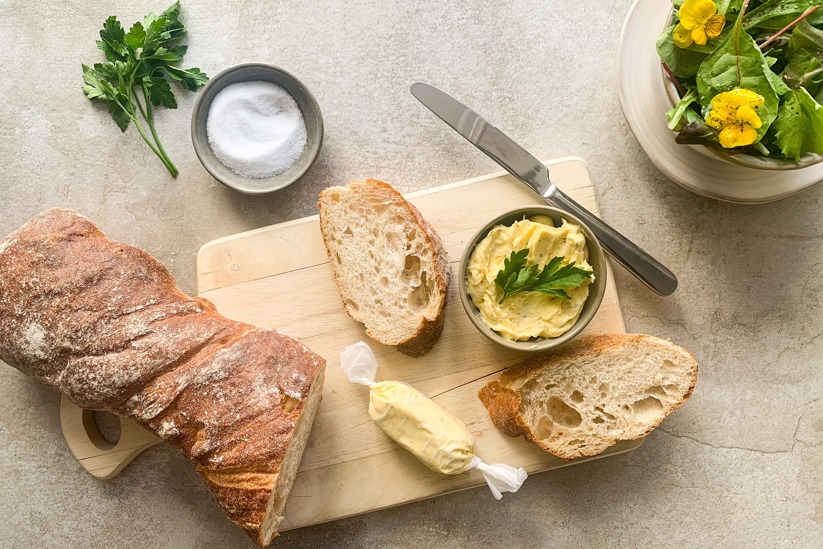 Кочан хляб върху дъска с две филийки и масло в купичка, отстрани - зелена салата.