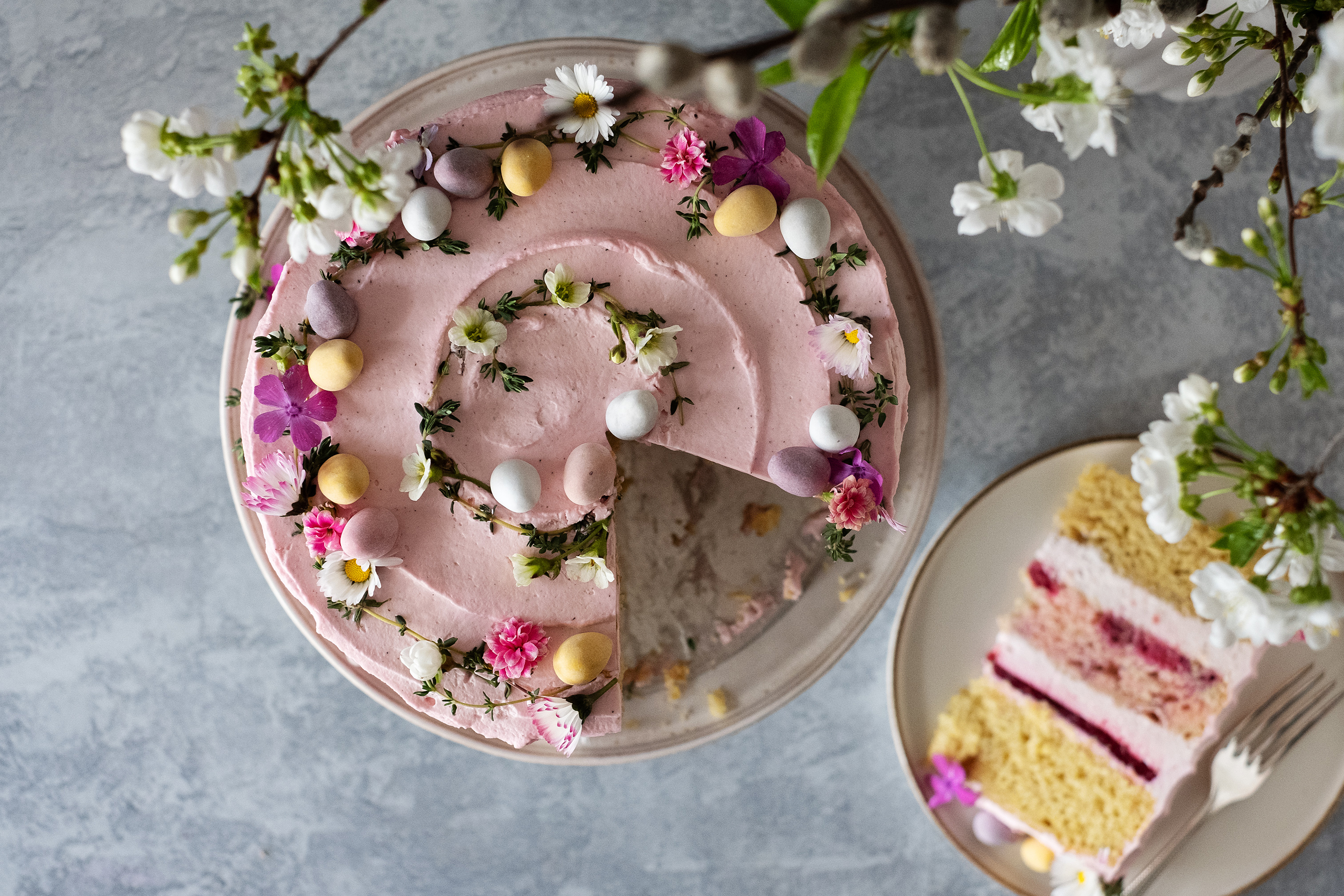 Торта с розова глазура, декорирана с великденски бонбони във формата на яйце и свежи цветчета, от която е отрязано парче, сервирано в чиния отдясно, отгоре стърчи цъфнал ябълков клон.