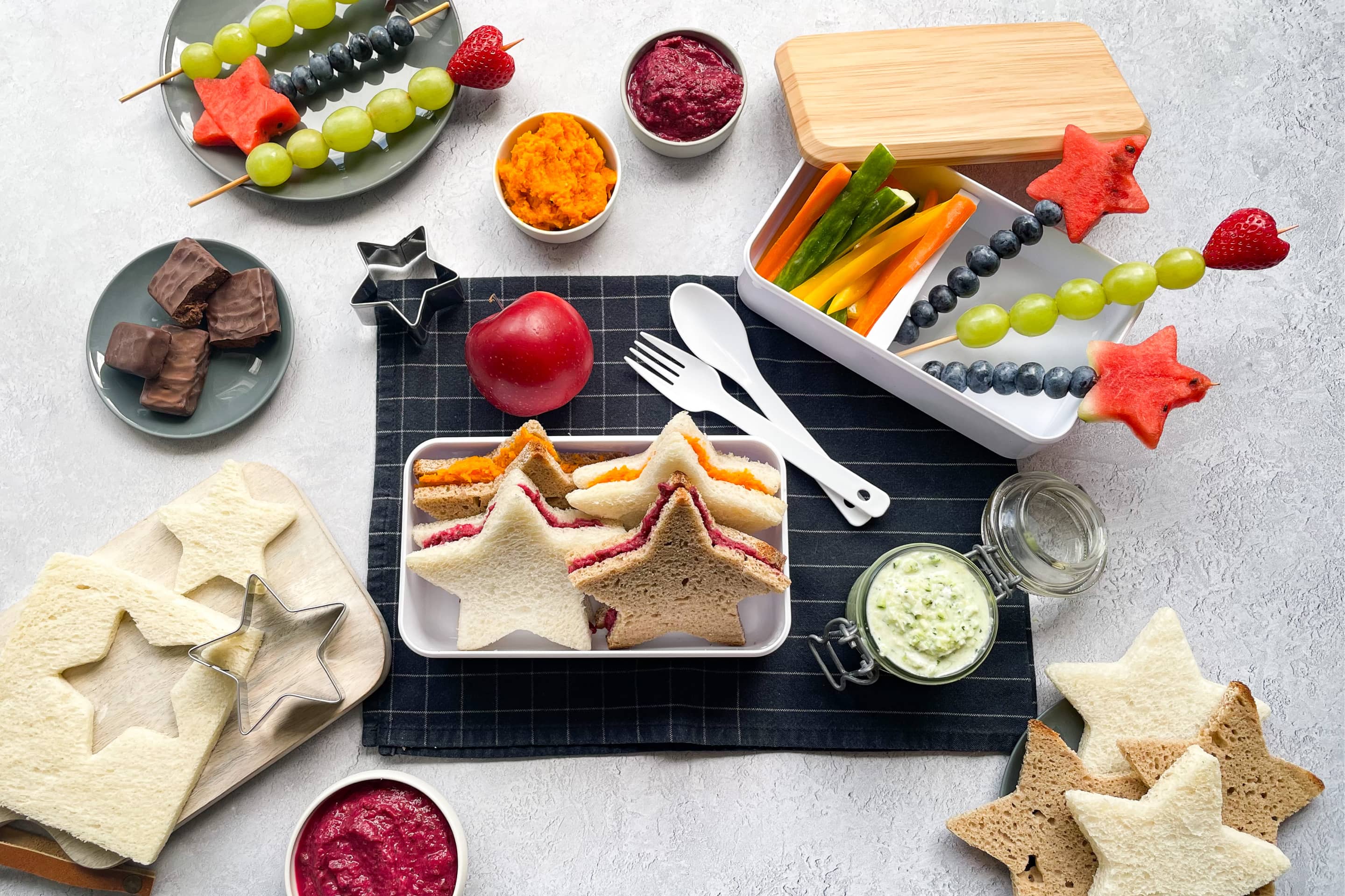 Сандвичи във формата на звездички в кутия за храна, намазани с цветни зеленчукови разядки и шишчета от плодове.