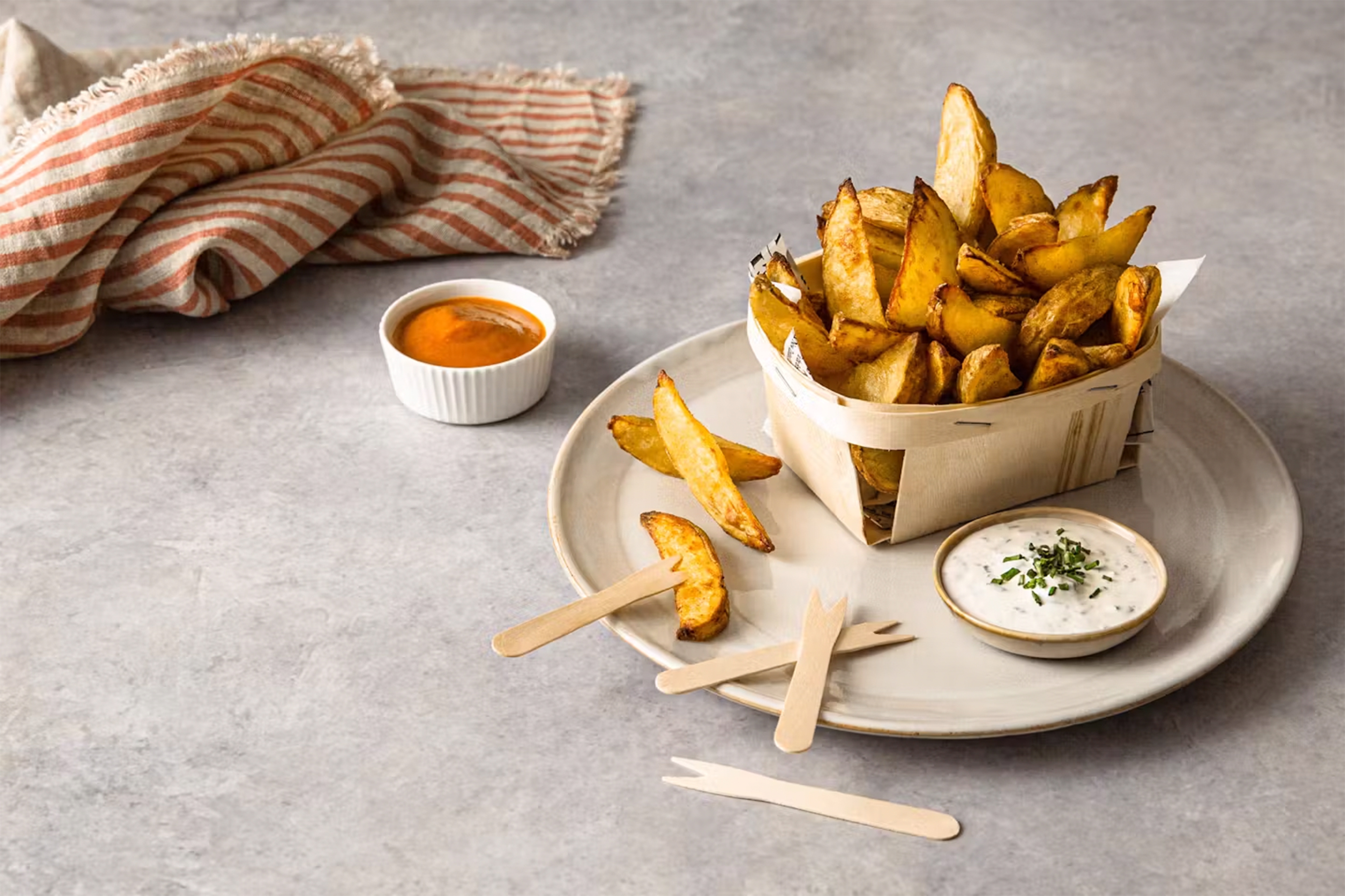Печени картофки в дървена кошничка, върху чиния с домашен кетчуп и млечен сос, поднесени в купички.