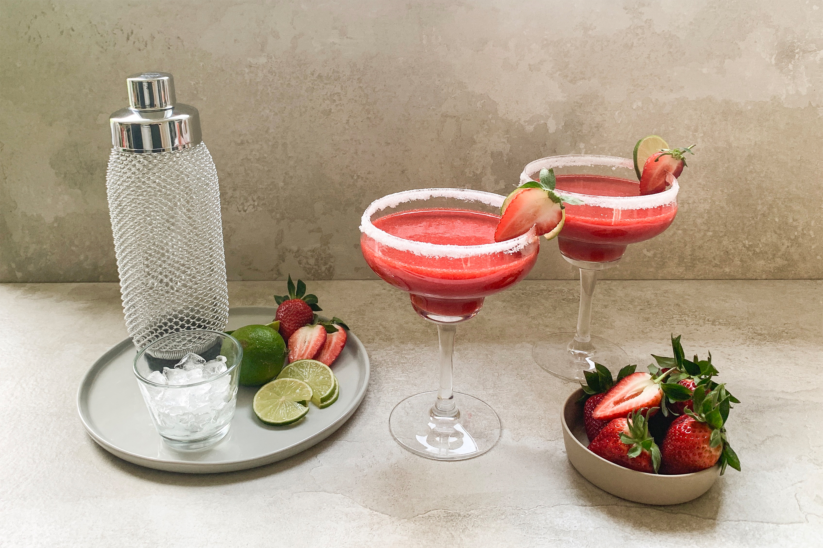 Две коктейлни чаши с ягодово дайкири, украсени с половинки ягода и захар по ръба, поднесени с купичка ягоди с листенцата и резенчета лайм.
